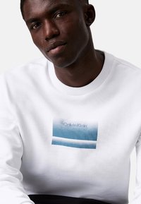 Sudadera blanca con un gráfico rectangular en degradado azul que presenta el texto "Calvin Klein". Cuello y puños acanalados, textura suave.