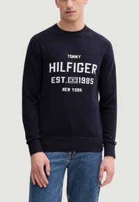 Granatowy sweter dziany z prążkowanym, okrągłym dekoltem, z białym nadrukiem tekstu "TOMMY HILFIGER EST. 1985 NEW YORK" z przodu.