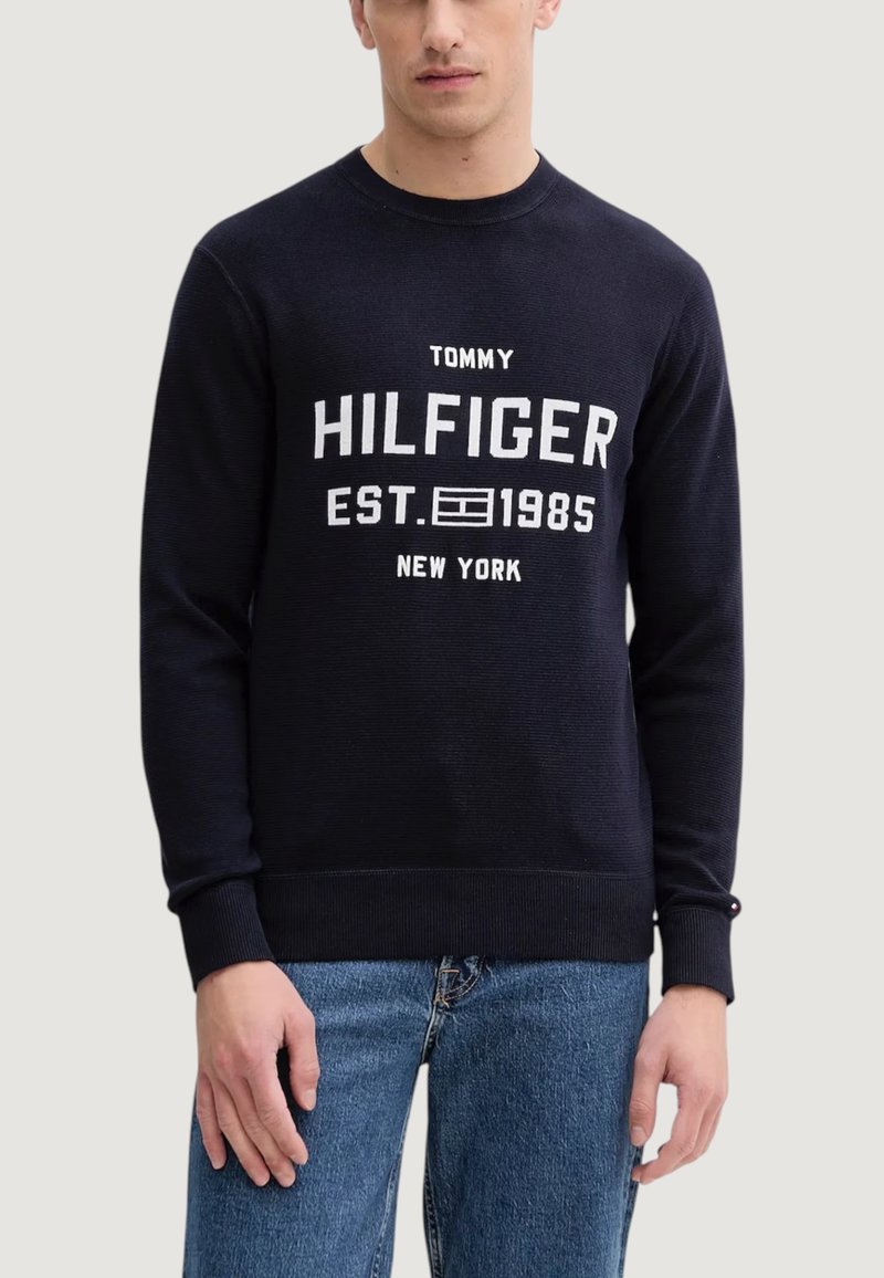 Granatowy sweter dziany z prążkowanym, okrągłym dekoltem, z białym nadrukiem tekstu "TOMMY HILFIGER EST. 1985 NEW YORK" z przodu.