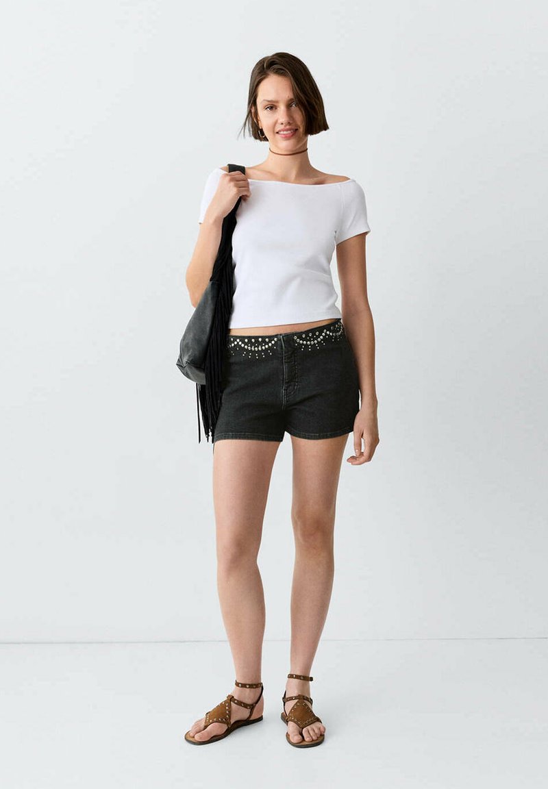 Jeune femme portant un haut blanc à manches courtes, un short noir clouté, des sandales marron cloutées et tenant un sac à épaule noir à franges sur fond uni.