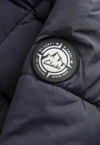 Sezione giacca a vento navy con un patch rotondo bianco e nero che presenta un design di montagna, coordinate e il testo "L'avventura inizia qui."