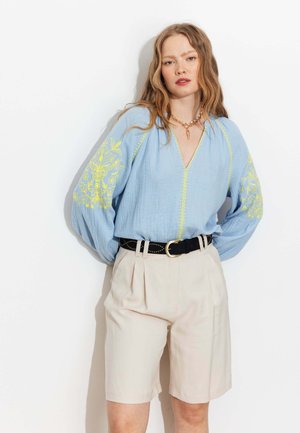 Femme portant un chemisier bleu clair avec broderies florales jaunes sur les manches, rentrée dans un short beige taille haute avec une ceinture noire, debout contre un mur blanc.