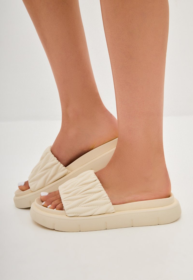 Sandales à glissière matelassées beige avec une semelle épaisse à finition texturée, un design ouvert à l'arrière et des détails de couture subtils sur les brides.