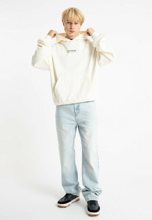 Biała bluza z kapturem i przednią kieszenią, zestawiona z jasnoniebieskimi dżinsami i czarnymi trampkami. Modelka pozuje z podniesionym kapturem.