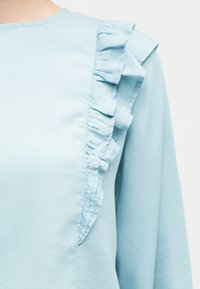 Ανοιχτό γαλάζιο πουκάμισο με μακριά μανίκια και βολάν κατά μήκος του μπροστινού και του ώμου, απλό φόντο, χωρίς ορατά λογότυπα ή σχέδια.