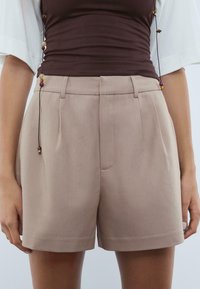 Close-up van een persoon die beige op de taille hoge tailleurschoenen en een bruine aansluitende top draagt, met een hanger van kralenketting naar beneden.