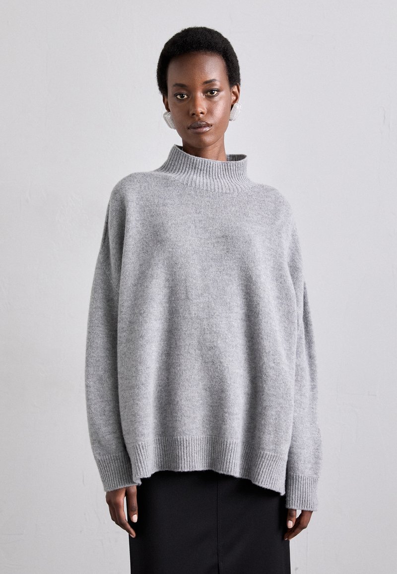 Pull en maille grise chunky avec un col montant côtelé, des épaules tombantes et des poignets et un ourlet côtelés. Texture douce et coupe décontractée.