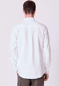 Camicia bianca a maniche lunghe con piega sul retro, orlo inferiore arrotondato e polsini abbottonati; tessuto leggero e texture liscia.