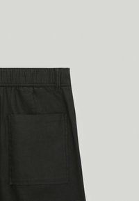 Pantalon noir avec une large ceinture élastique, comprenant une seule poche arrière et un tissu lisse et texturé. Le design est simple et fonctionnel.