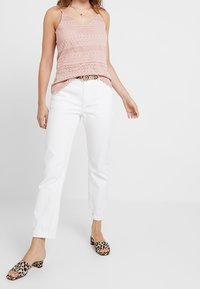 Růžový krajkový top s výstřihem do V, bílé džíny s rovným střihem a nazouvací boty s leopardím vzorem. Má jemný vzor a nastavitelné ramínka.