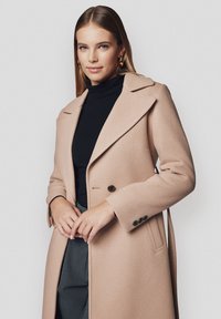 Beżowy wełniany płaszcz z klapami, z jednoszrowym zapięciem i trzema czarnymi guzikami. Nosi się go na czarnym golfie, charakteryzuje się eleganckim krojem.