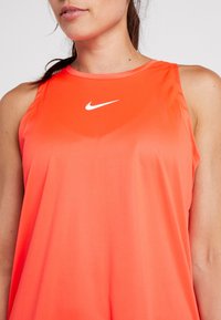 Femme portant un haut de sport Nike sans manches de couleur orange vif avec un logo Nike swoosh blanc sur la poitrine, vue du cou jusqu'au torse.