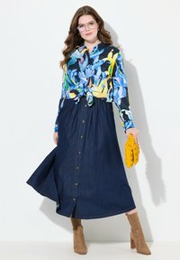 Blouse à manches longues florale avec un devant noué, associée à une jupe maxi en denim avec des boutons sur le devant et des fentes sur les côtés, accessoirisée d'une pochette jaune.