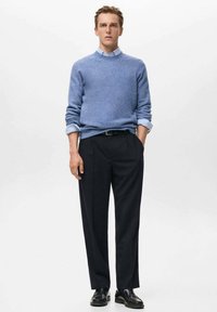 Bărbat în picioare, purtând un pulover bleu deschis peste o cămașă cu guler, pantaloni negri și mocasini negri, cu mâinile în buzunare, pe un fundal neutru.