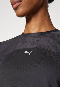 Camisa atlética negra con un patrón oscuro texturizado en los hombros y un logo de Puma blanco en el pecho. Tejido suave y diseño ajustado.