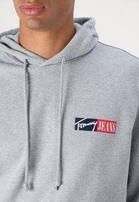 Grå hættetrøje lavet af en blanding af bomuldsstof, med en forlommem og et logo patch med "Tommy JEANS" i rød og marineblå.