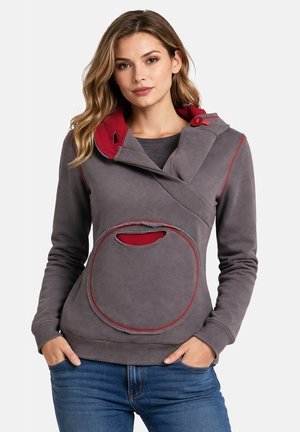 Donna che indossa una felpa con cappuccio in pile grigia e rossa con colletto regolabile e tasca frontale circolare, abbinata a jeans blu.