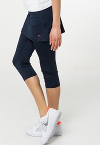 Falda corta de atletismo de color navy sobre leggins capri negros, hecha de un tejido elástico. Zapatillas deportivas blancas con detalles en naranja completan el look.