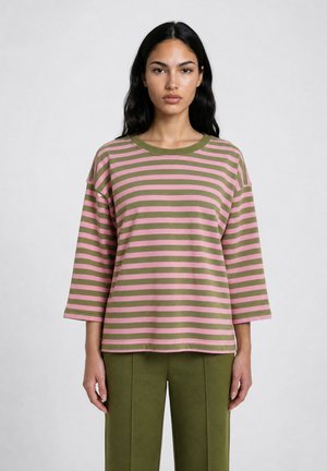 Woman with long black hair wearing a pink and green striped long-sleeve shirt and olive green pants, standing against a plain light background.