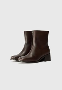 Bottes en cuir marron, à cheville, avec un bout arrondi, une fermeture éclair sur le côté et un talon carré ; texture lisse et design minimaliste.