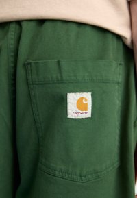 Žalios medvilninės kelnės turi užpakalinę kišenę su balta etikete, ant kurios pavaizduotas geltonas logotipas ir tekstas "carhartt". Minkšta, patvari medžiaga su tvarkingu dizainu.