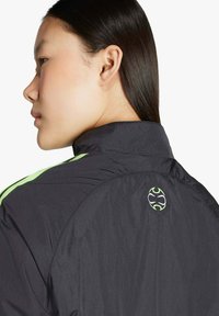 Junge Frau mit langen, glatten Haaren trägt eine schwarze Jacke mit neongrünen Streifen und einem kleinen runden Logo am Rückenkragen.