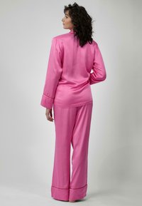 Pigiama di raso rosa con maniche lunghe e pantaloni. La parte superiore ha un colletto e polsini, mentre i pantaloni presentano una gamba ampia e dettagli sul bordo.