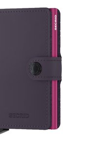 Portafoglio in pelle viola con un accento rosa. Presenta una forma arrotondata, bordi cuciti e una chiusura a scatto in metallo con un dettaglio logo impresso.