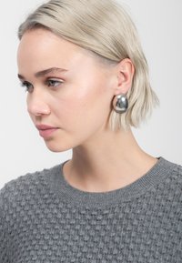 Jonge vrouw met kort blond haar, die grote zilveren ovale oorbellen en een gestructureerde grijze trui draagt, en nadenkend naar haar links kijkt.