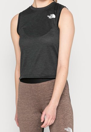 Nainen pukeutunut hihattomaan mustaan North Face -toppiin ja ruskeisiin North Face -legginsseihin, seisoo vaalean yksinkertaisen taustan edessä.