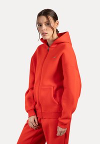 Rode rits hoodie van dik fleecemateriaal, met een capuchon, voorzakken en geribde manchetten. Subtiele logo op de borst, relaxed fit.
