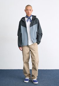 Lightweight, two-tone windbreaker in gray and light blue, with a zip front and side pockets. Worn over a white T-shirt and beige pants.