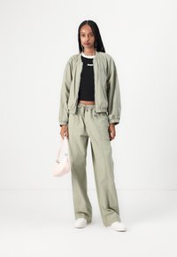 Ljusgrön bomberjacka med elastiska ärmar, matchande lösa byxor och en svart crop top. Håller en liten rosa handväska. Vita sneakers.