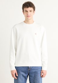Sudadera blanca de algodón con mangas largas, puños y bajo acanalados, presenta un pequeño logo rojo en el pecho. Combinada con jeans de mezclilla azul.
