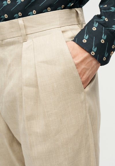 Χέρι στην τσέπη μπεζ λινών παντελονιών που φοριούνται με σκούρο πουκάμισο με μικρό λουλουδάτο σχέδιο και μανσέτα με κουμπιά.