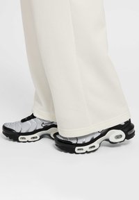 Sneakers Nike z czarną podeszwą, szarością w górnej części z falistym wzorem i białym obramowaniem. W zestawieniu z beżowymi spodniami, które zakrywają część butów.