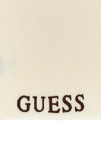 Tejido de canalé crema con una textura de punto, que presenta letras en marrón oscuro "GUESS" destacadas cerca de la parte inferior.