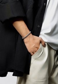 Braccialetto in argento indossato su un polso, con anelli intrecciati. La mano è infilata in una tasca di pantaloni beige chiaro, in contrasto con una camicia scura.