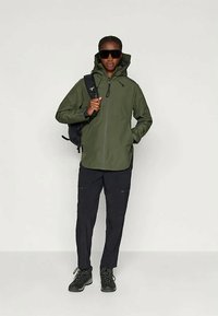 Osoba s tmavými slnečnými okuliarmi, zelenou kapucňou, čiernymi nohavicami, turistickými topánkami a čiernym ruksakom stojí proti svetlému pozadiu.