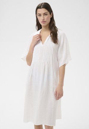 Ung kvinde med langt brunt hår iført en løs, hvid knælængde kjole med plisseret front og brede ærmer til albuerne, står på hvid baggrund.