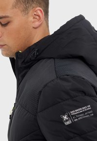 Černá nadýchaná bunda s texturovaným šestiúhelníkovým vzorem na ramene, vybavená kapucí, zapínáním na zip a logem na rukávu. Minimaliustní design.