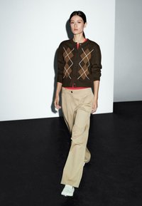 Frau, die drinnen geht, trägt einen braunen Argyle-Pullover, ein rotes Hemd, beigefarbene weite Hosen und weiße Schuhe auf einem schwarz-weißen Boden.