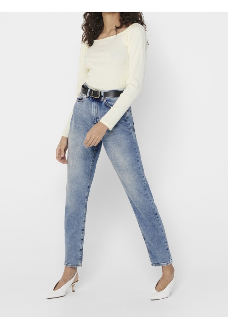 Femme portant un haut à manches longues jaune clair, un jean bleu délavé taille haute avec une ceinture noire, et des chaussures blanches à talons bas, debout sur un fond blanc.