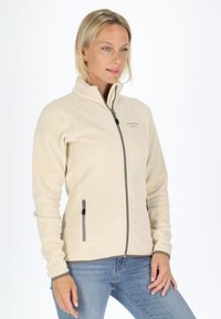 Lichtbeige fleece jack met hoge kraag, ritssluiting aan de voorkant en twee zijzakken; bevat grijze ritsen en subtiele branding op de borst.