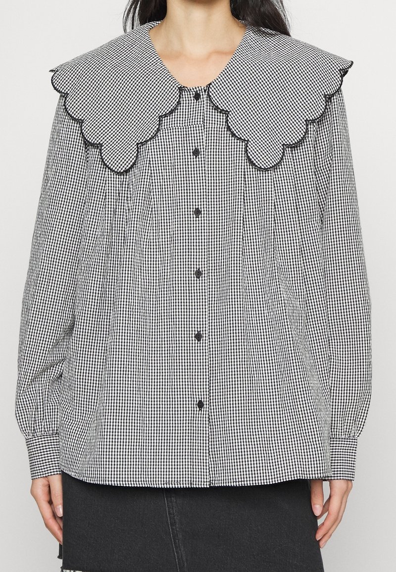 Blouse vichy noire et blanche avec grand col festonné, boutons noirs sur le devant et manches longues bouffantes avec poignets boutonnés.