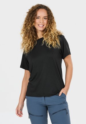 Lächelnde Frau mit lockigem Haar, die ein schwarzes Kurzarmshirt und blaue Hosen mit Reißverschlusstaschen trägt, steht mit einer Hand in der Tasche.