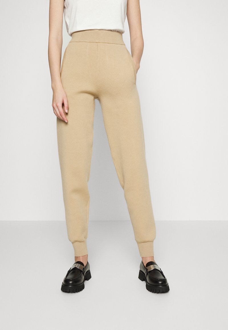Personne portant un pantalon beige en maille taille haute avec des chevilles retroussées et des mocassins noirs épais, debout devant un fond blanc uni.
