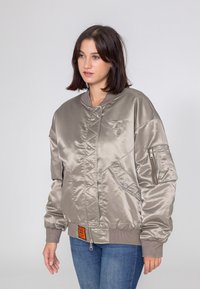 Srebrna satynowa kurtka bomberka z zamykanym na zamek kieszonką, ściągaczami przy mankietach i logo na piersi. Posiada gładką teksturę i pikowaną konstrukcję.