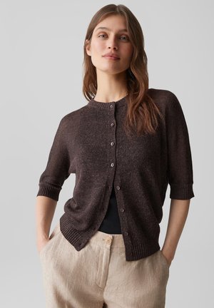 Jonge vrouw met bruin haar die een donkerbruine, geknoopte cardigan draagt over een zwart topje en beige broek met handen in de zakken.