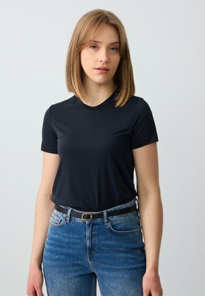 Schwarzes T-Shirt mit kurzen Ärmeln aus weichem Stoff, mit einem runden Halsausschnitt. Kombiniert mit hochgeschnittenen, ausgewaschenen blauen Jeans und einem schwarzen Gürtel.